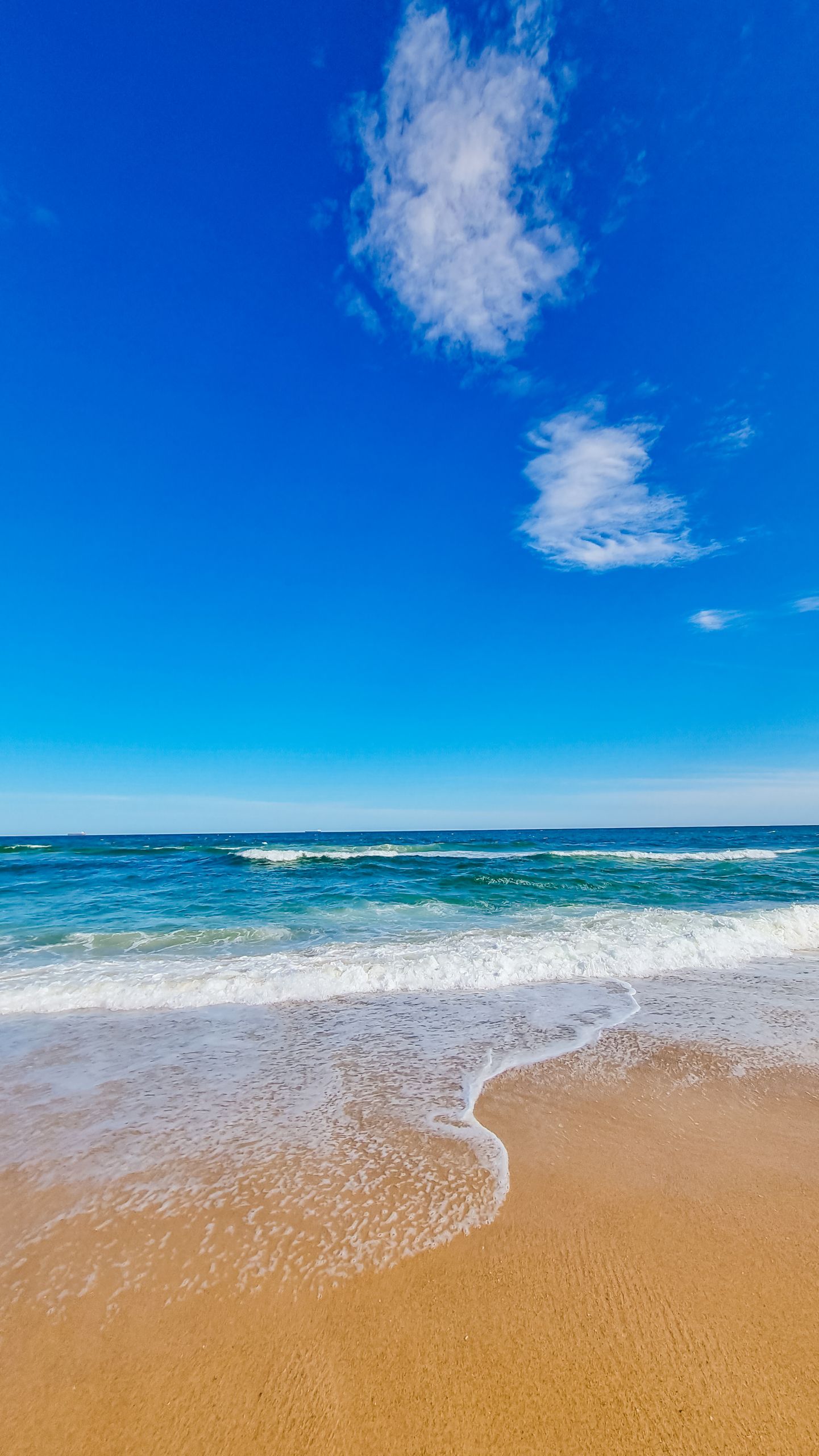 Serene scene of ocean waves crashing on a sandy beach under a vibrant blue sky.